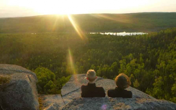 two students rest against rocks overlooking a vast green forest containing a body of water